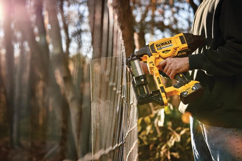 Une personne utilise une agrafeuse sans fil DEWALT pour fixer une clôture en fil dehors, avec des arbres et de la lumière du soleil en arrière-plan.