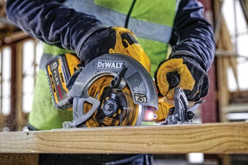 Vue rapprochée d'une personne utilisant une scie circulaire sans fil DEWALT 60V FLEXVOLT pour couper un morceau de bois. La personne porte des gants de travail et un gilet haute visibilité à l'intérieur d'un chantier de construction.