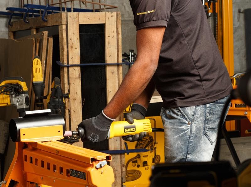Personne utilise une meuleuse DEWALT DWE4887N sur métal dans un atelier avec d'autres outils jaunes.