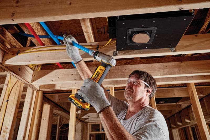 Une personne portant des gants utilise un outil sans fil DEWALT pour travailler sur un tuyau bleu dans un chantier à ossature bois.