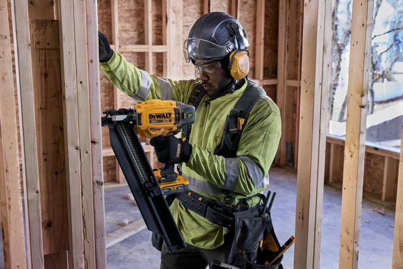 Un ouvrier du bâtiment portant des équipements de sécurité utilise un cloueur DEWALT DCN920B pour fixer l'ossature en bois à l'intérieur d'un bâtiment en construction. Le visage de l'ouvrier est flouté.