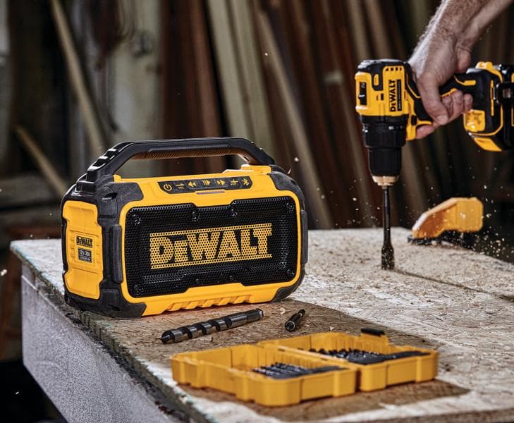 Une enceinte Bluetooth portable DEWALT posée sur un établi à côté d'une perceuse sans fil DEWALT en cours d'utilisation, des forets et une boîte d'organisation jaune, dans un atelier.