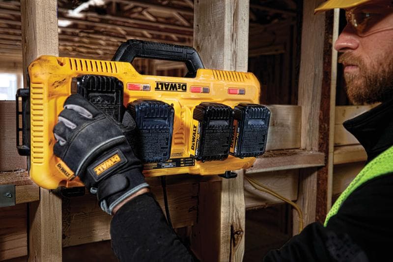 Une personne portant des gants de travail installe ou retire des batteries d'un chargeur multi-batteries jaune DEWALT fixé sur une poutre en bois sur un chantier de construction.