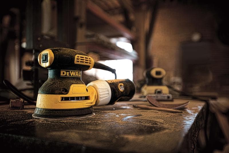 Gros plan sur une ponceuse électrique DEWALT DWE6421 placée sur un établi poussiéreux dans un atelier, avec des pads de ponçage et une autre ponceuse visibles à l'arrière-plan.