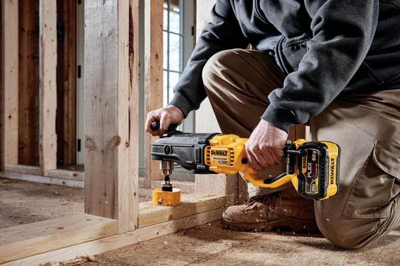 A person is kneeling on a construction site using a DEWALT DCD470X1 cordless drill with a hole saw attachment to drill through a wooden stud frame.