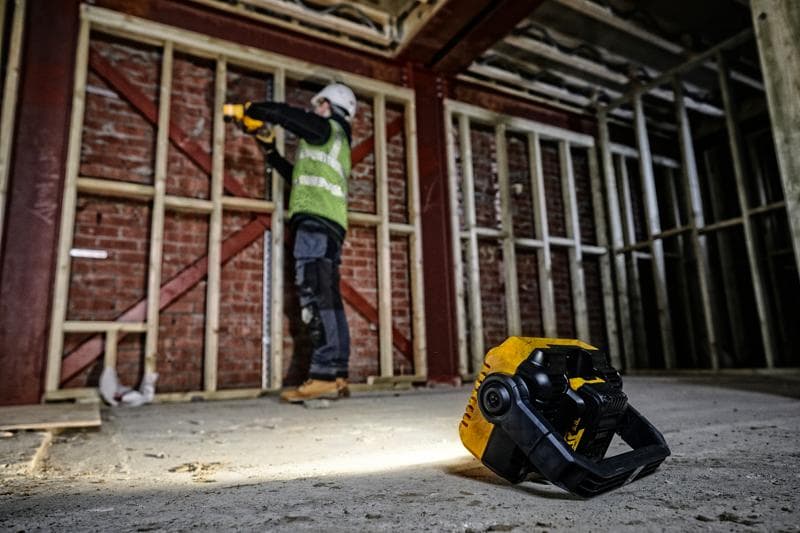 Lampe de chantier DEWALT DCL077 au sol, ouvrier en arrière-plan près d'une structure en bois.
