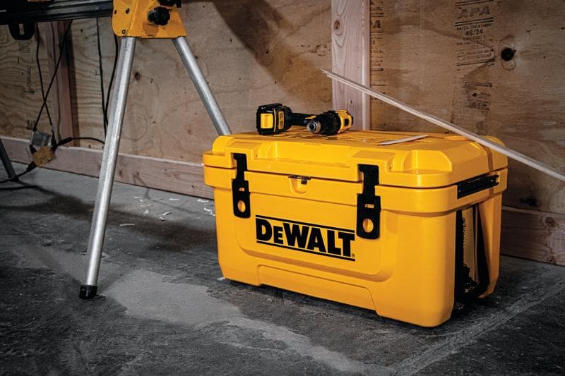 Une boîte à outils jaune DEWALT posée sur un sol en béton, avec une perceuse électrique reposant dessus. La boîte à outils est située près de pieds métalliques et d'un mur en bois dans un atelier.