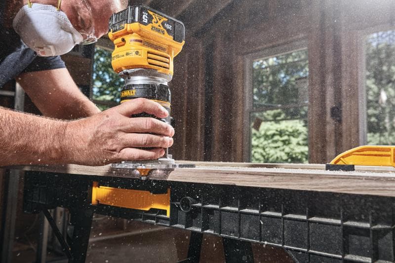 Gros plan sur une personne utilisant une défonceuse sans fil DEWALT pour façonner le bord d'une planche en bois sur un établi, avec de la sciure volante et la personne portant un masque de protection.