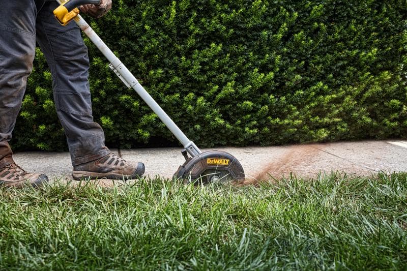 Une personne utilise un coupe-bordure DEWALT pour tailler le bord d'une pelouse près d'un trottoir, avec des buissons verts en arrière-plan.