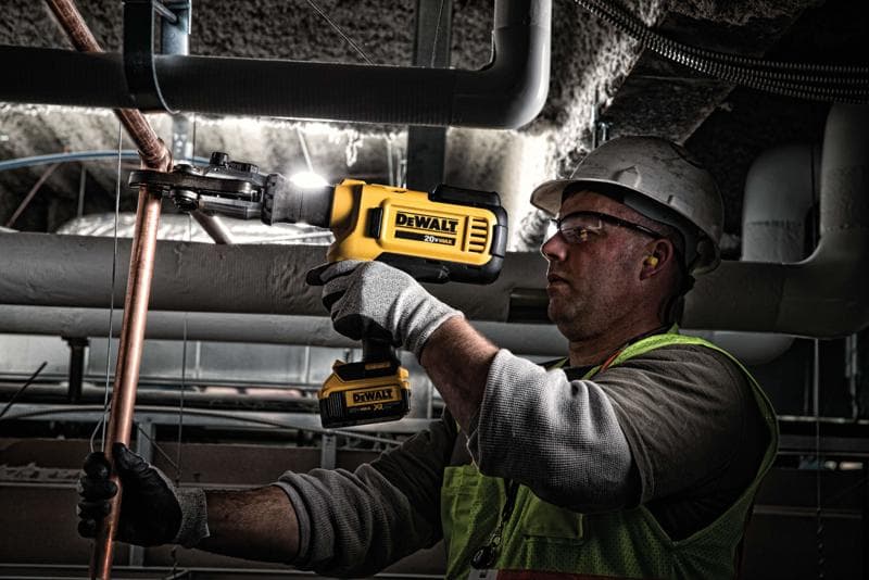 Une personne portant un équipement de sécurité utilise un outil sans fil jaune DEWALT pour couper ou façonner un tuyau en cuivre dans un environnement industriel.