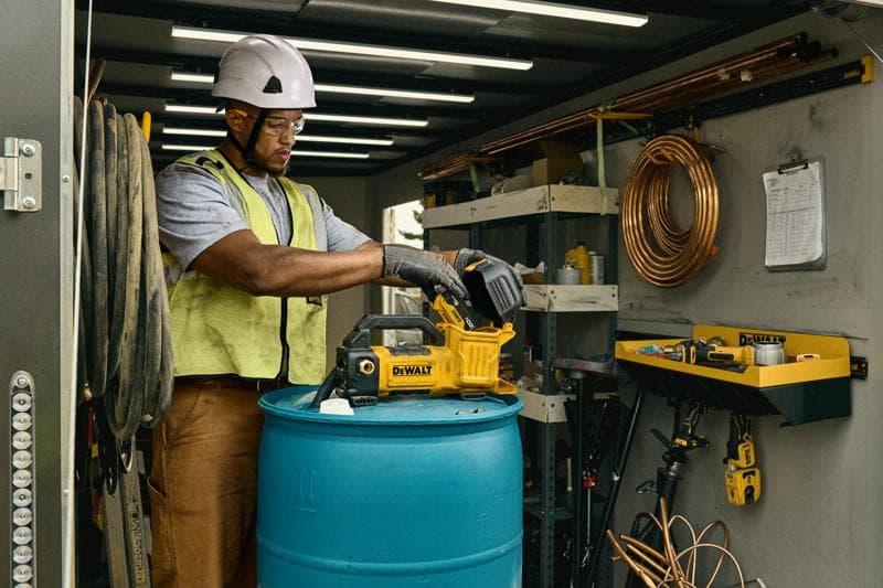 Une personne portant un casque de sécurité et un gilet utilise un outil DEWALT, modèle DCE050P1, sur un tonneau bleu à l'intérieur d'un atelier ou d'un espace de stockage. L'espace comporte des étagères, des tuyaux en cuivre enroulés et d'autres outils et équipements visibles.