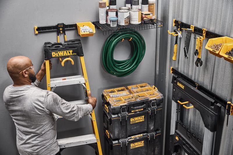 Une personne organise un espace de travail avec des produits de rangement DEWALT, notamment une échelle, des boîtes à outils, des bacs muraux, des outils à main, un tuyau d'arrosage et des étagères avec des pots de peinture.