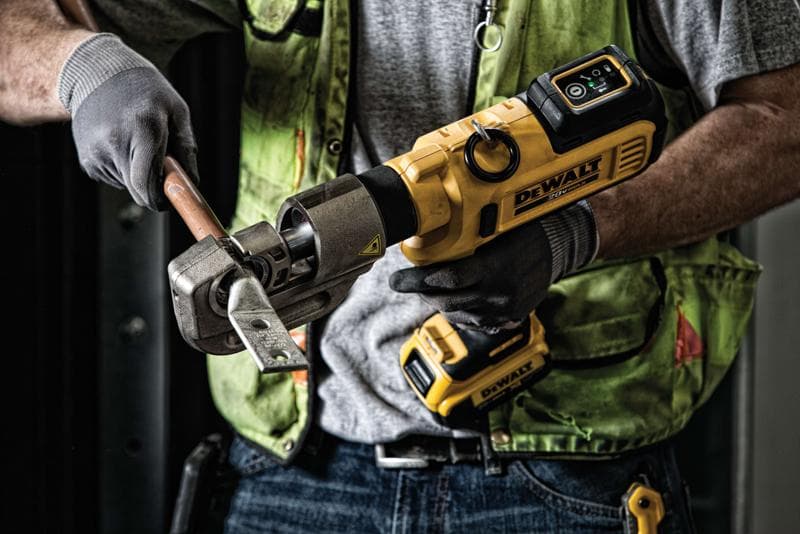Une personne utilise un outil sans fil DEWALT pour couper une tige métallique épaisse tout en portant des gants de travail et un gilet de sécurité vert.