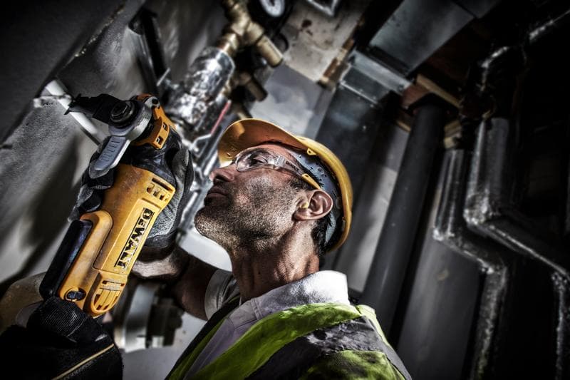Ouvrier coupe un tuyau mural avec un DEWALT DWE315, équipé d'un casque et d'un gilet dans un espace industriel.