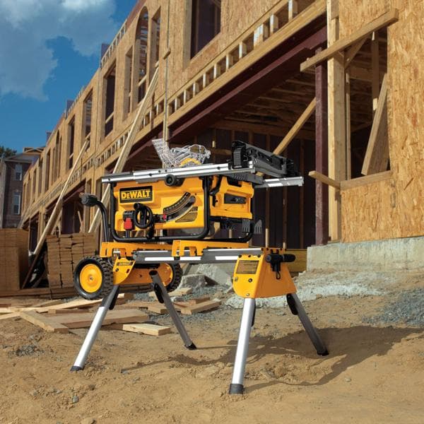 Une scie sur table portable DEWALT avec un support est présentée sur un chantier de construction, avec un bâtiment en bois partiellement construit en arrière-plan. La scie et son support sont posés sur un sol sablonneux, avec des matériaux de construction tels que des planches en bois empilées à proximité.