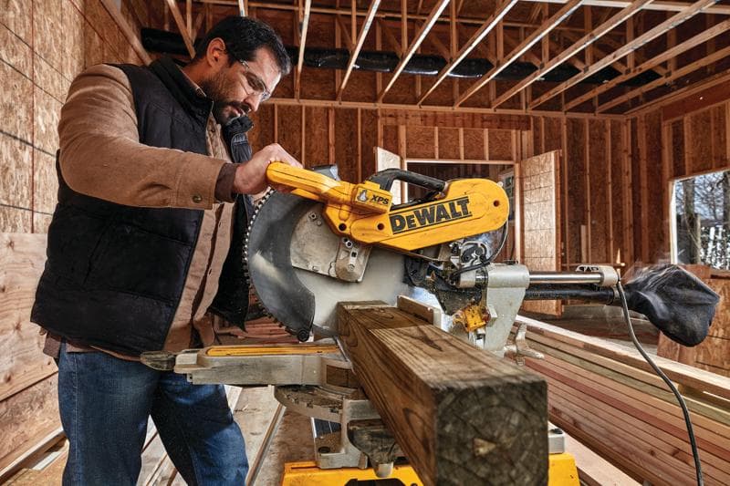 Une personne utilise une scie à onglets DEWALT DWS780 pour couper une poutre en bois sur un chantier de construction. La scie est jaune et porte le logo DEWALT de façon bien visible. L’arrière-plan montre une ossature en bois exposée et des murs non finis.