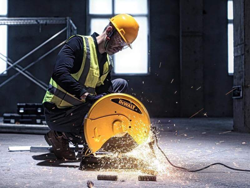 Une personne portant un casque de sécurité jaune et un gilet réfléchissant utilise un outil de coupe DEWALT D28730 pour couper des tuyaux en métal dans un environnement industriel, avec des étincelles visibles provenant de la machine.