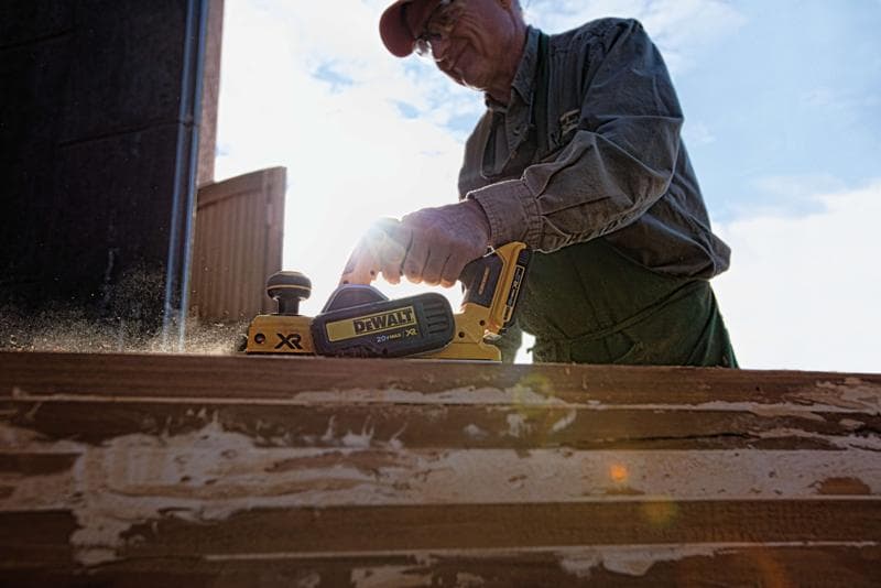 Une personne utilisant une raboteuse sans fil DEWALT DCP580B 20V sur une surface en bois à l’extérieur, avec des copeaux de bois projetés lors de l’utilisation de la raboteuse.