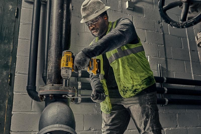Un ouvrier portant un casque et un gilet de sécurité utilise un outil électrique jaune DEWALT pour fixer des boulons sur un gros tuyau industriel à l'intérieur d'une installation.
