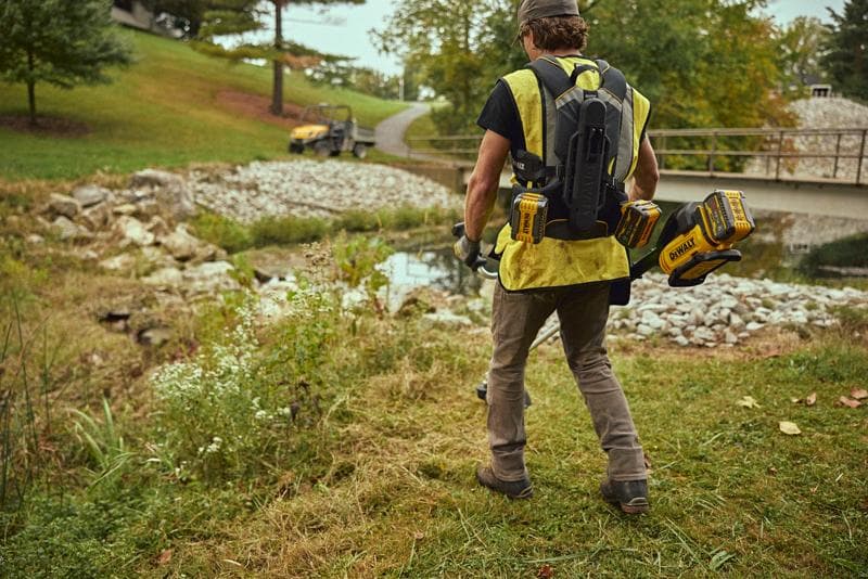 Une personne portant un gilet jaune et un système de batterie sac à dos DEWALT marche à l'extérieur près d'un ruisseau, avec des batteries d'outils électriques DEWALT attachées au sac à dos.