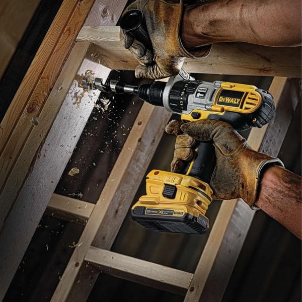 Une personne portant des gants de travail marron utilise une perceuse électrique sans fil DEWALT pour percer un trou dans une poutre en bois, avec des copeaux de bois projetés par le processus de perçage.