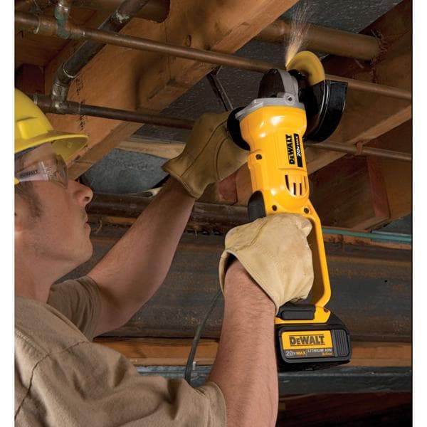 Une personne portant des gants de protection utilise une meuleuse d’angle sans fil DEWALT jaune pour couper un tuyau en métal sous un plafond en bois. La meuleuse fonctionne sur batterie et produit des étincelles lors de la coupe.