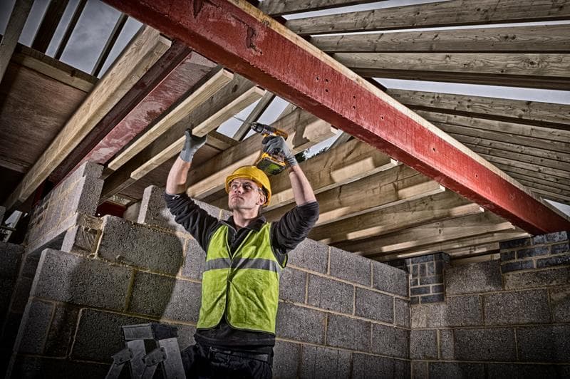 Un ouvrier du bâtiment portant un casque de sécurité jaune et un gilet haute visibilité utilise une perceuse sans fil DEWALT pour fixer des poutres en bois à une poutre d'acier rouge dans un bâtiment en construction. Le visage de l'ouvrier est flouté.