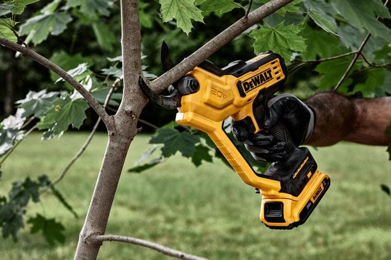 Une personne portant un gant noir utilise un sécateur électrique sans fil DEWALT 20V jaune pour couper une branche d’arbre à l’extérieur.