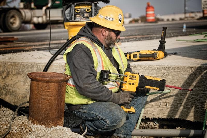Un ouvrier du bâtiment portant un casque jaune, un gilet de sécurité et des gants utilise un outil électrique DEWALT pour percer du béton sur un chantier extérieur. Le visage de l’ouvrier est flouté pour préserver sa confidentialité. D'autres outils DEWALT et du matériel de construction sont visibles à l’arrière-plan.
