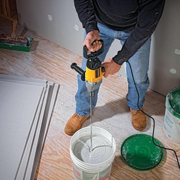 A person using a DEWALT DW130V power mixer to mix paint or plaster in a large bucket on a wooden floor. Drywall sheets and construction materials are visible nearby.