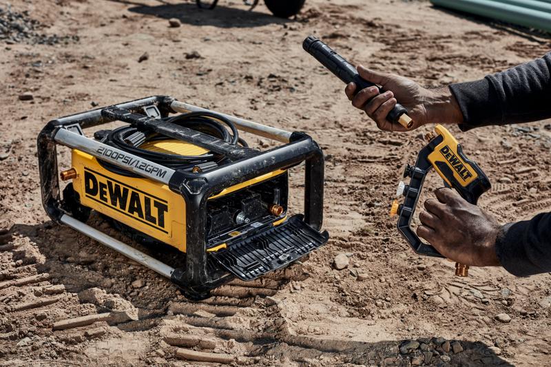 Un nettoyeur haute pression portable DEWALT avec des tuyaux et accessoires visibles est en cours d'assemblage à l'extérieur sur une surface en terre. Une personne tient une buse de pulvérisation et la connecte à la machine.