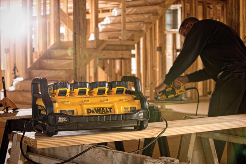 Une station d'alimentation portable DEWALT avec quatre emplacements pour batteries est placée sur un établi dans un chantier de construction. Une personne en arrière-plan utilise un outil électrique sur une planche en bois.