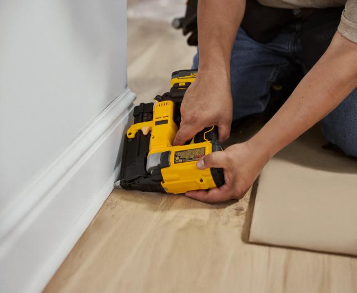 Une personne agenouillée au sol utilise une cloueuse sans fil jaune DEWALT pour fixer une plinthe au mur dans un intérieur de maison.