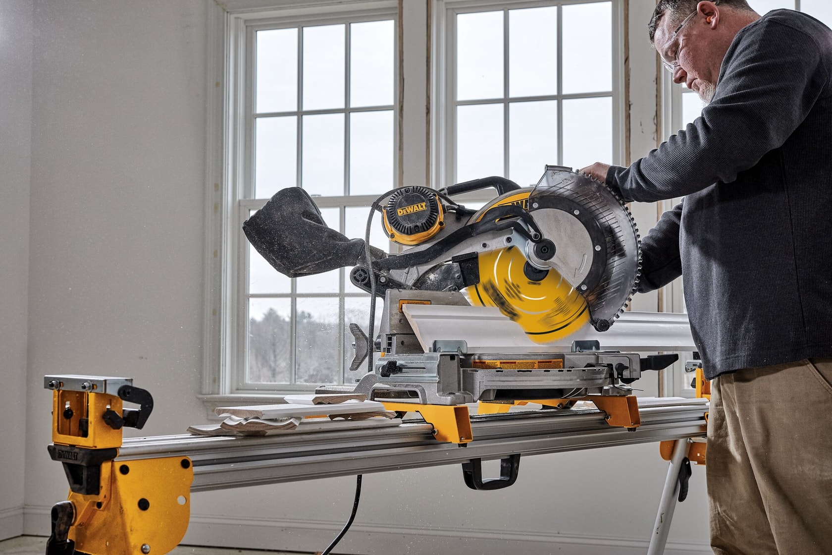 Une personne utilise une scie à onglet DEWALT pour couper une moulure en bois dans une pièce lumineuse, avec la scie placée sur un support jaune et argenté. Des fenêtres sont visibles en arrière-plan et des travaux de menuiserie sont en cours.