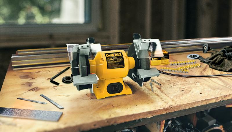 Une meuleuse d'établi DEWALT, modèle DW758, est posée sur un établi en bois. Divers outils et pièces métalliques l'entourent, suggérant un espace de travail actif. La meuleuse est jaune et possède deux roues à meuler avec des carters de sécurité.