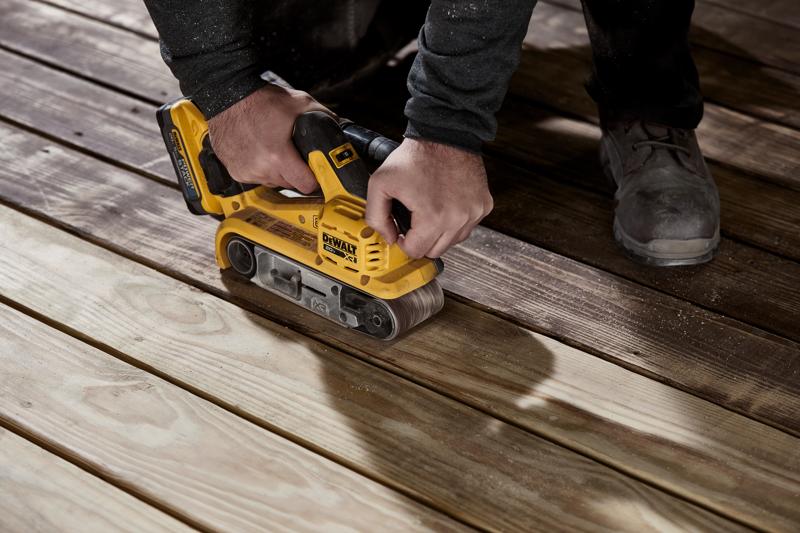 Une personne utilise une ponceuse à bande sans fil jaune DEWALT pour poncer des planches en bois sur un sol.
