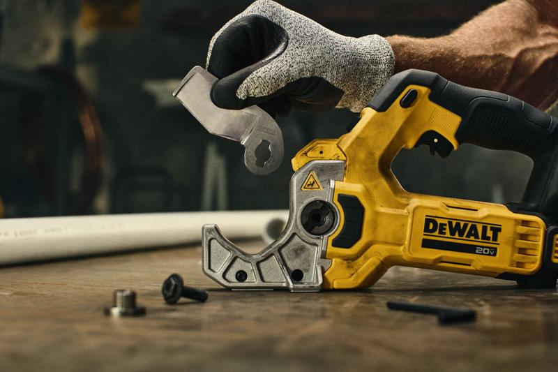 Une personne portant un gant de protection tient un accessoire de lame à côté d'un outil électrique jaune DEWALT 20V, placé sur un établi. De petites pièces de matériel sont visibles sur la table et un tuyau en plastique blanc se trouve à l'arrière-plan.