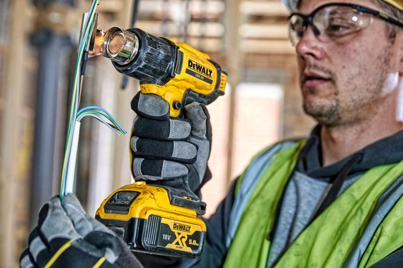 Une personne portant des gants de travail et un gilet de sécurité utilise un pistolet à chaleur sans fil DEWALT DCE530N pour appliquer de la chaleur sur des fils électriques sur un chantier de construction. Le visage de la personne est flouté pour préserver la confidentialité.