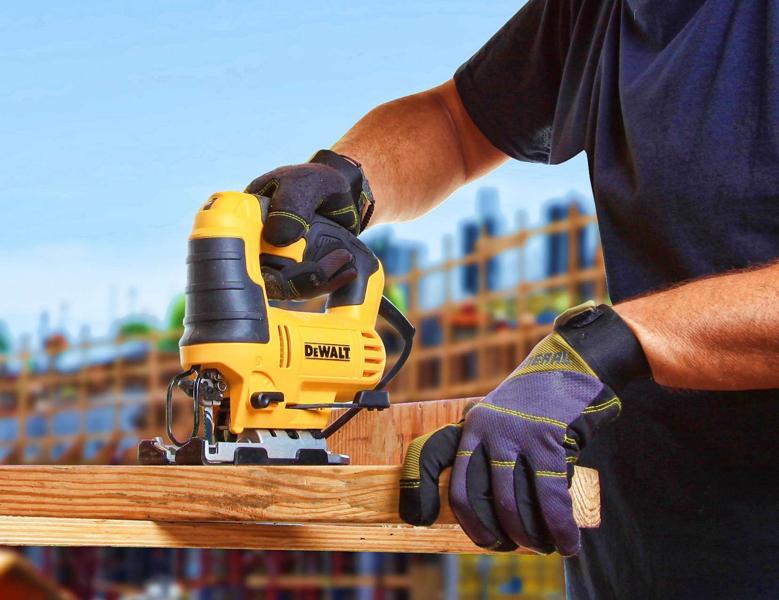 Une personne portant des gants de protection utilise une scie sauteuse jaune DEWALT pour couper une pièce de bois, avec un chantier visible en arrière-plan.