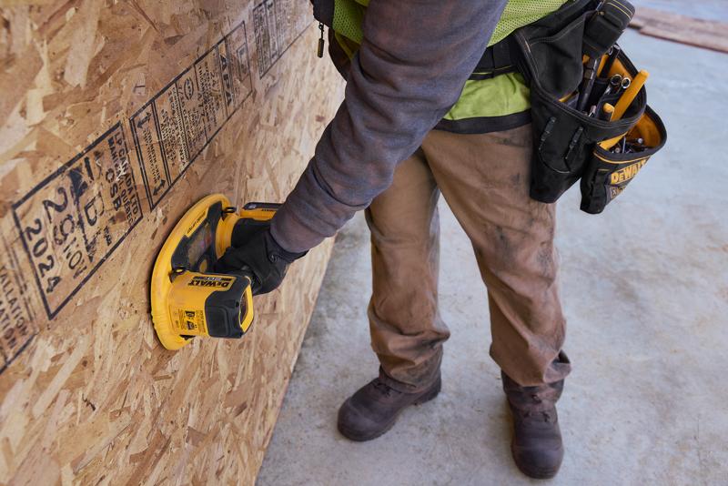 Un travailleur portant un pantalon marron et des gants de protection utilise un outil portatif DEWALT contre une grande plaque d'OSB sur un chantier de construction. Le travailleur porte une ceinture à outils avec divers outils à main.