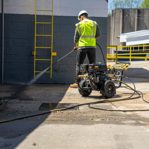 A person in safety gear operates a DEWALT pressure washer to clean a concrete surface outdoors near a building with a yellow ladder.