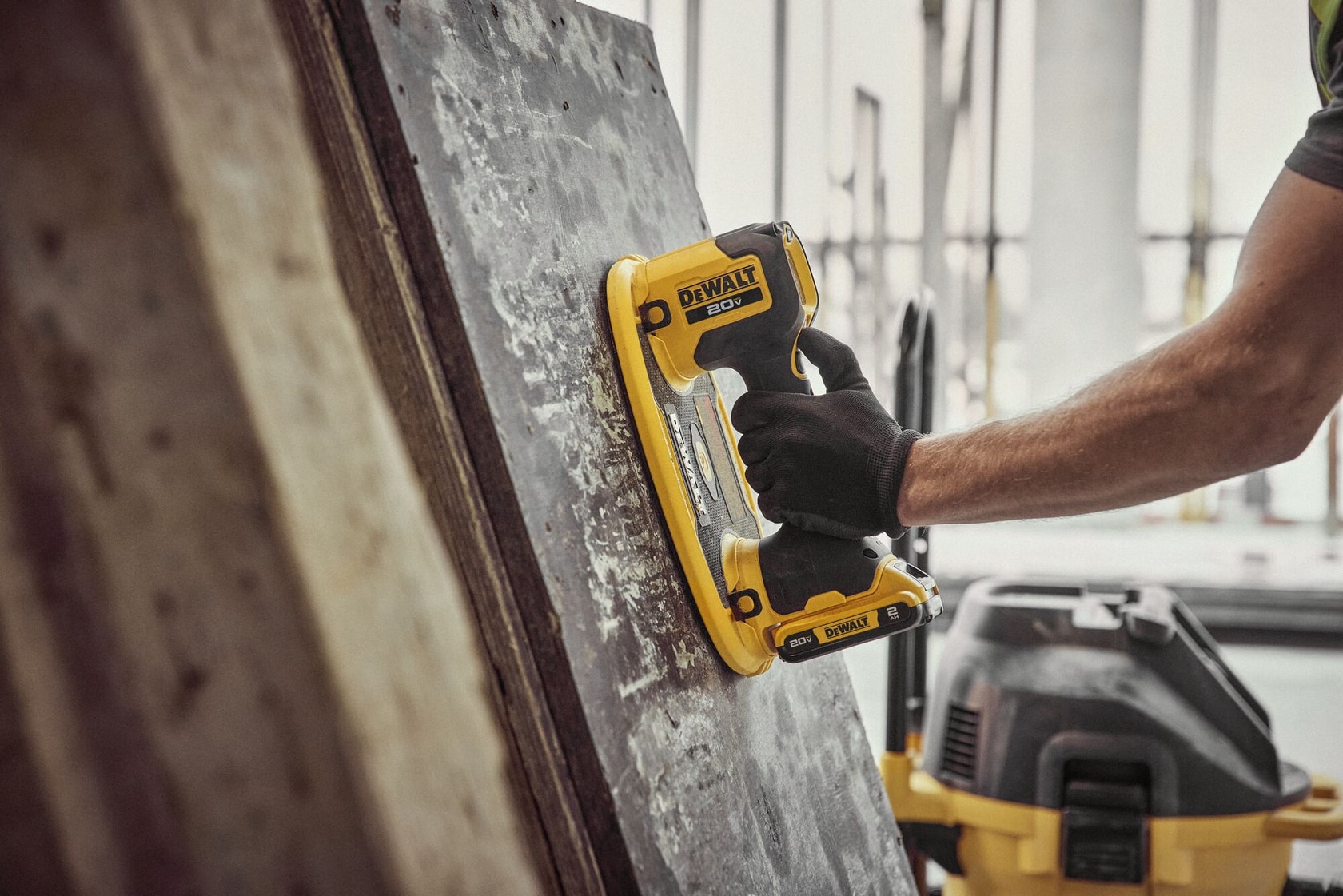 Une main gantée utilise une ponceuse portative sans fil DEWALT 20V sur une grande surface en bois usée, avec un autre outil DEWALT en arrière-plan.