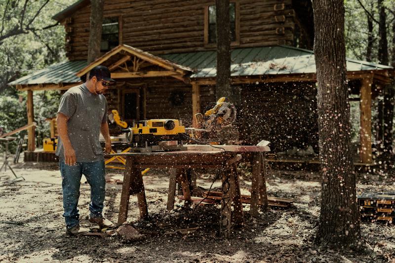 Une personne utilise une scie à onglet DEWALT DWS780 à l'extérieur, coupant du bois sur un établi devant une cabane en rondins. De la sciure vole dans l'air tandis que la personne opère l'outil. L'environnement comprend des arbres et du matériel de menuiserie.