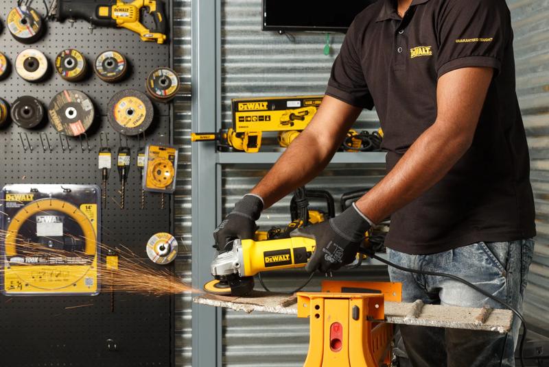 Une personne portant des gants utilise une meuleuse angulaire Dewalt DW803 pour couper une pièce métallique, provoquant des étincelles. Divers outils électriques Dewalt et disques de meulage sont présentés sur un mur et un établi en arrière-plan.