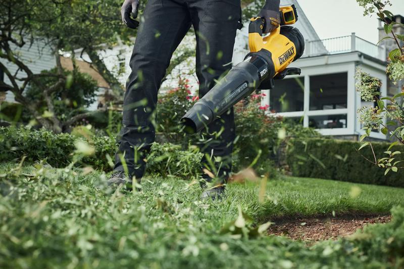 Person using a yellow and black DEWALT cordless leaf blower to clear grass clippings and leaves in a garden or yard.