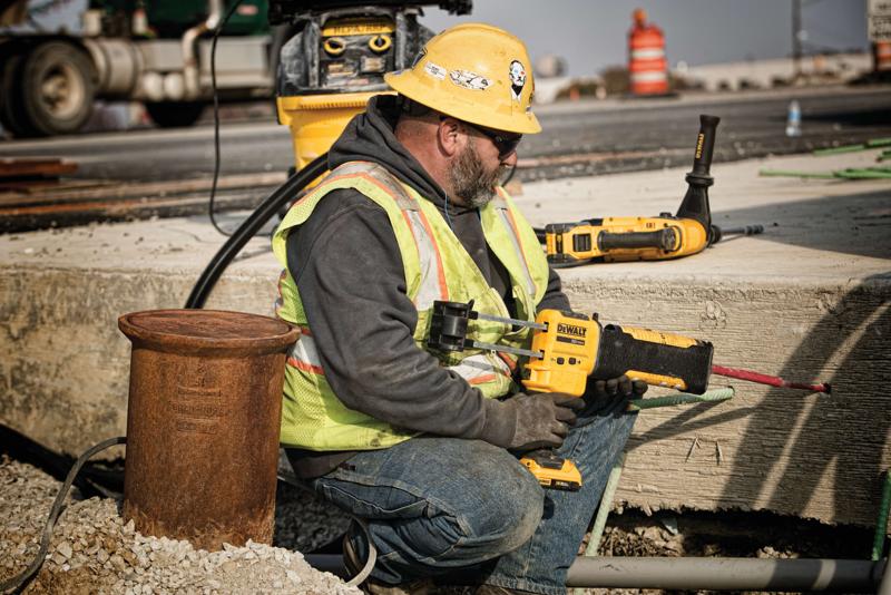 Un ouvrier du bâtiment portant un casque jaune, un gilet de sécurité et des gants utilise un outil électrique DEWALT pour percer du béton sur un chantier extérieur. Le visage de l’ouvrier est flouté pour préserver sa confidentialité. D'autres outils DEWALT et du matériel de construction sont visibles à l’arrière-plan.