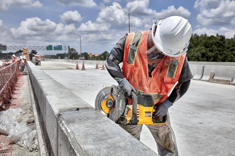 Un ouvrier du bâtiment portant un casque blanc et un gilet de sécurité orange utilise une scie sans fil DEWALT (modèle DCS692B) pour couper une barrière en béton sur un chantier routier.