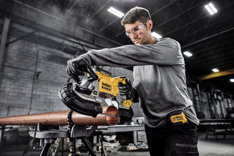 Une personne utilise une scie à ruban portable DEWALT DCS374B pour couper un tuyau métallique dans un atelier industriel. Le visage de la personne est flouté pour des raisons de confidentialité.