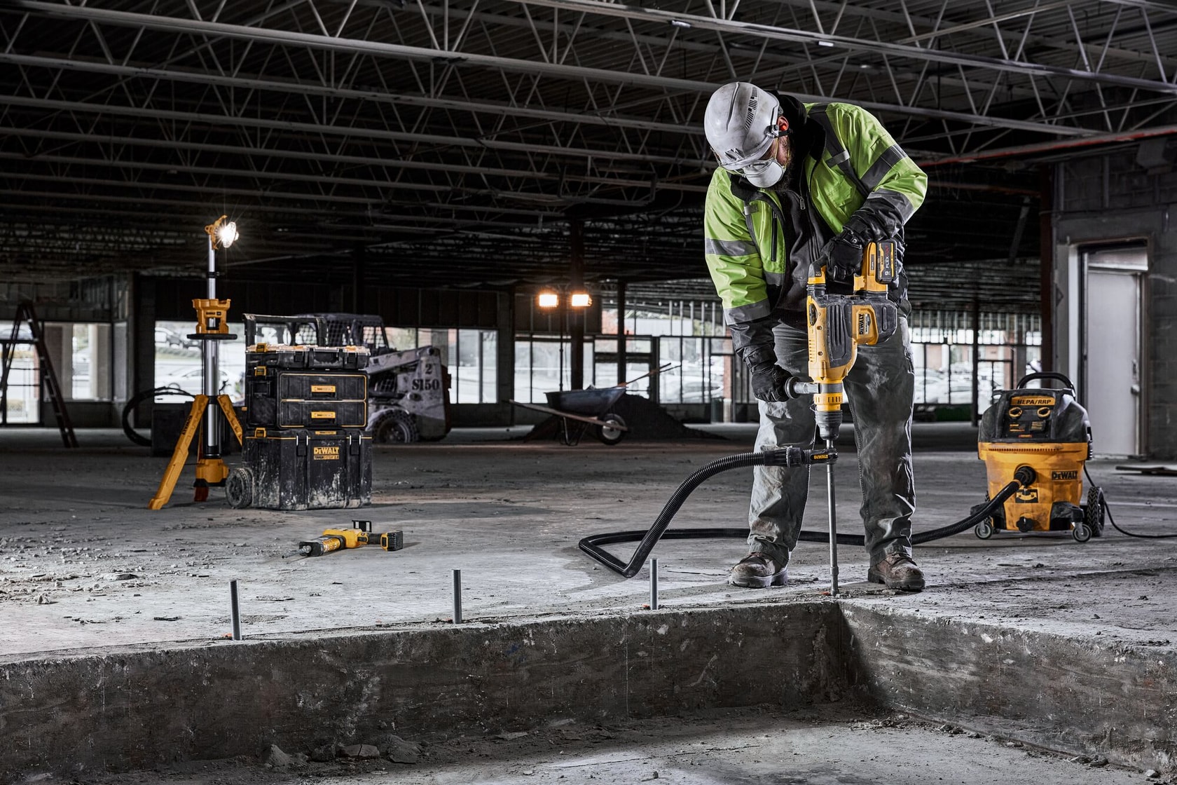 Un ouvrier du bâtiment portant un équipement de sécurité utilise un outil électrique DEWALT pour percer du béton à l'intérieur d'un bâtiment en construction. Divers outils et équipements DEWALT sont visibles autour de la zone de travail.